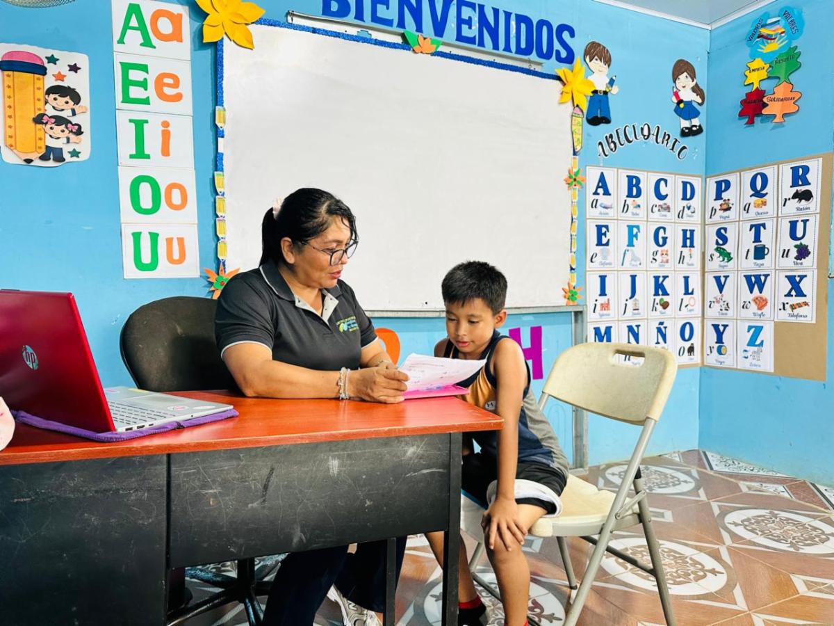 One-on-one tutoring: a female teacher sits at a red desk helping a young student with papers in a colorful classroom.