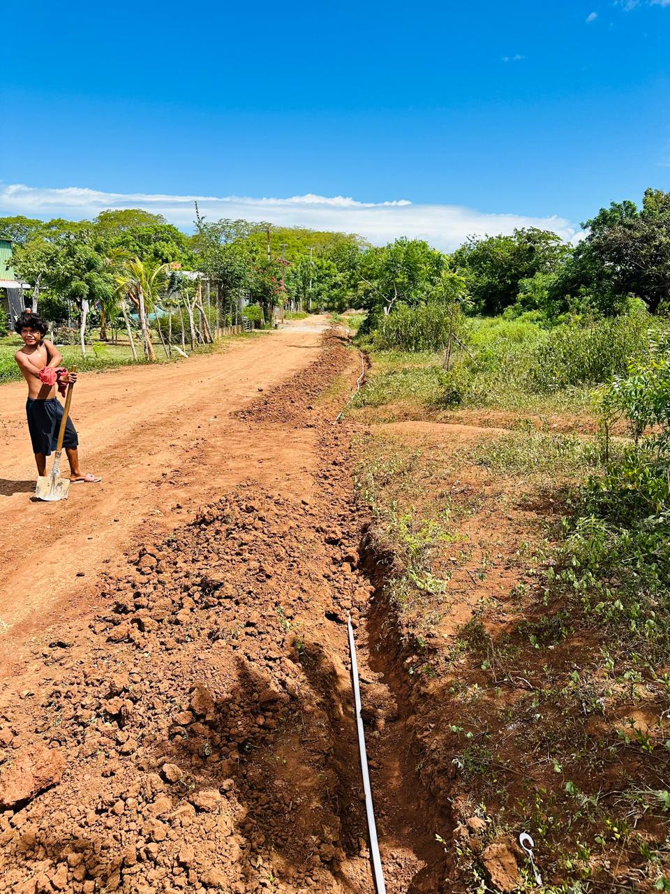 Boy with a shovel stands on a dusty rural road as a trench for a water pipe is dug along the path under a bright blue sky.