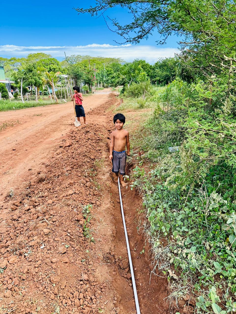 Two boys on a dusty rural road beside a deep trench laid with a long plastic pipe; one shirtless boy stands in the trench while another in a red shirt works farther back amid green vegetation.