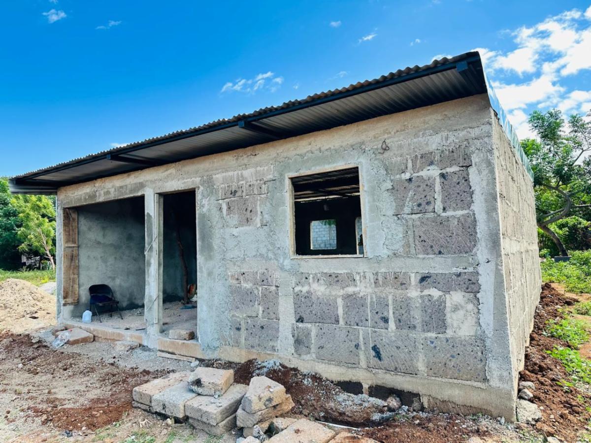 Unfinished concrete block building with a metal roof, open doorway and a small window, surrounded by construction rubble.