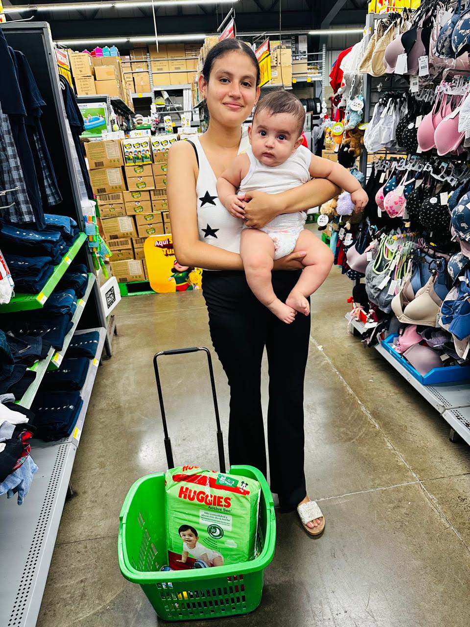 Woman in a white tank top holding a baby in a store aisle, with a green shopping basket in front containing a pack of Huggies diapers.