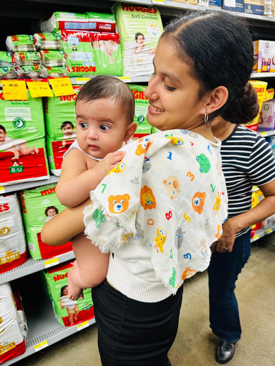 Woman holding a baby in an aisle of a store, looking at stacked diaper packs on shelves behind them