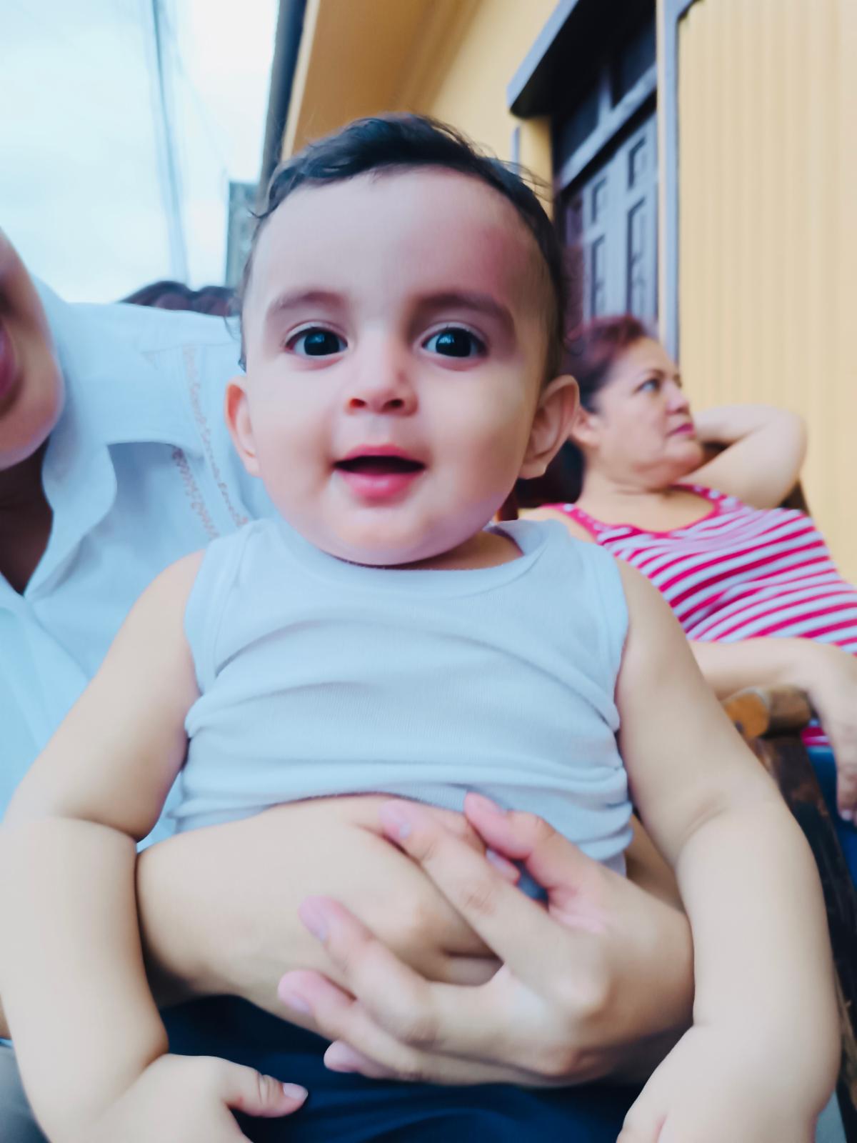 Baby smiling at the camera, held by adults outdoors; wearing a white sleeveless shirt.