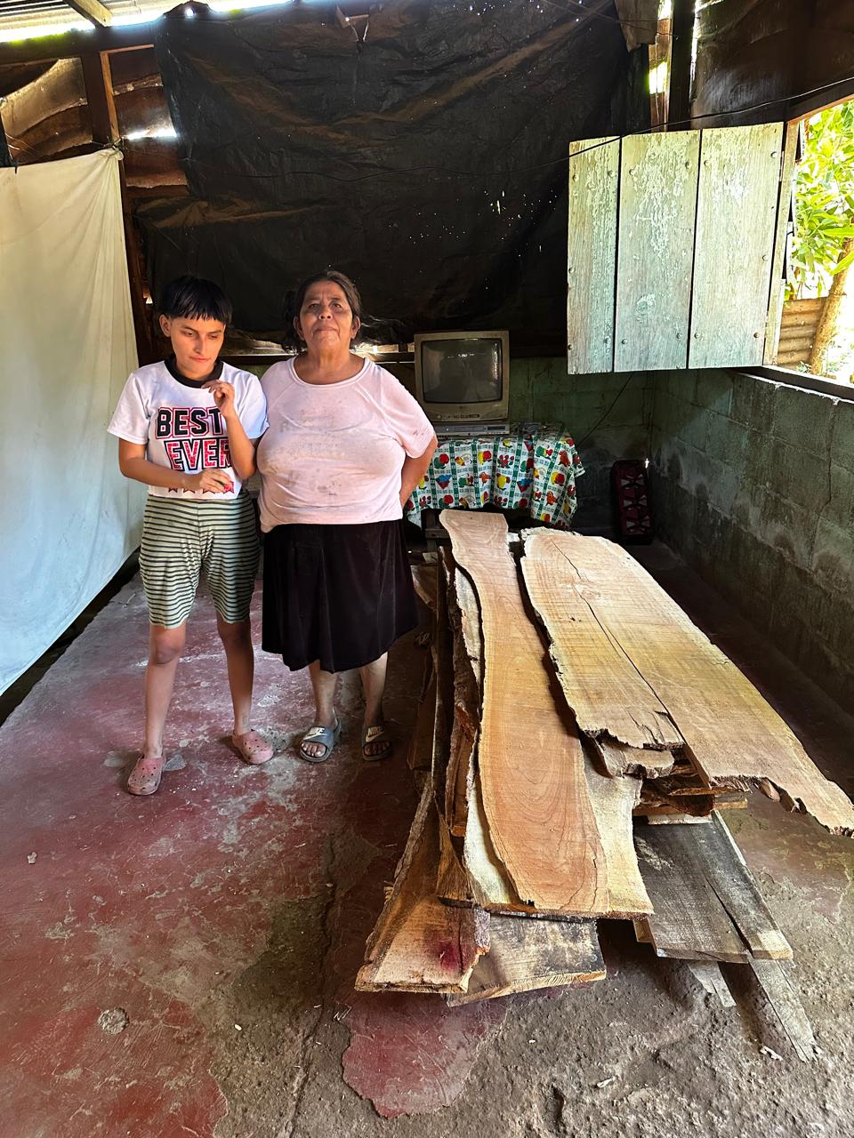 Boy and an older woman stand in a rustic woodworking shed beside rough-cut planks and a small old TV on a table covered with a colorful cloth.