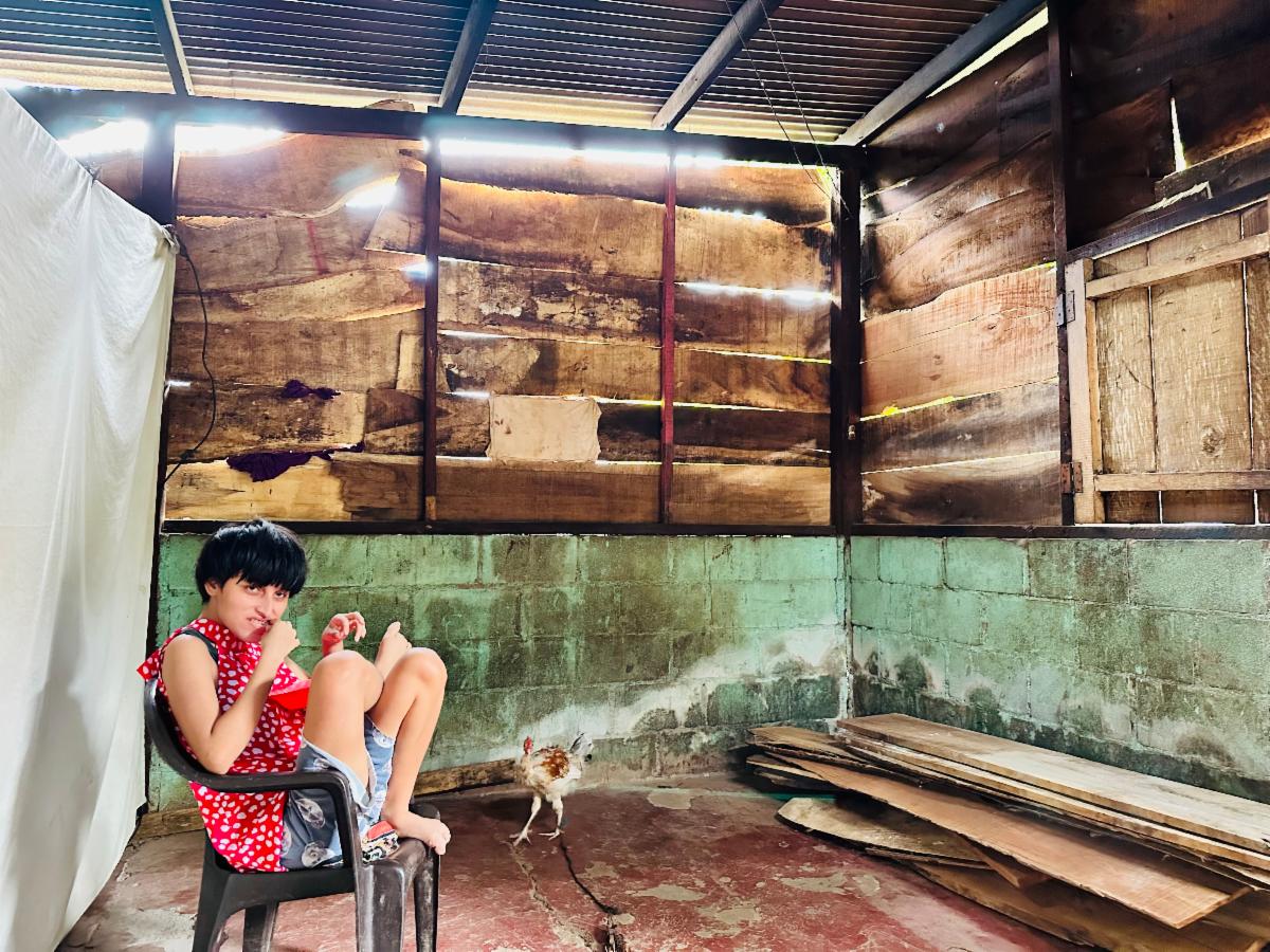 Girl in a red polka dot shirt sits in a plastic chair inside a rough wooden shed, a chicken nearby on the dirt floor.