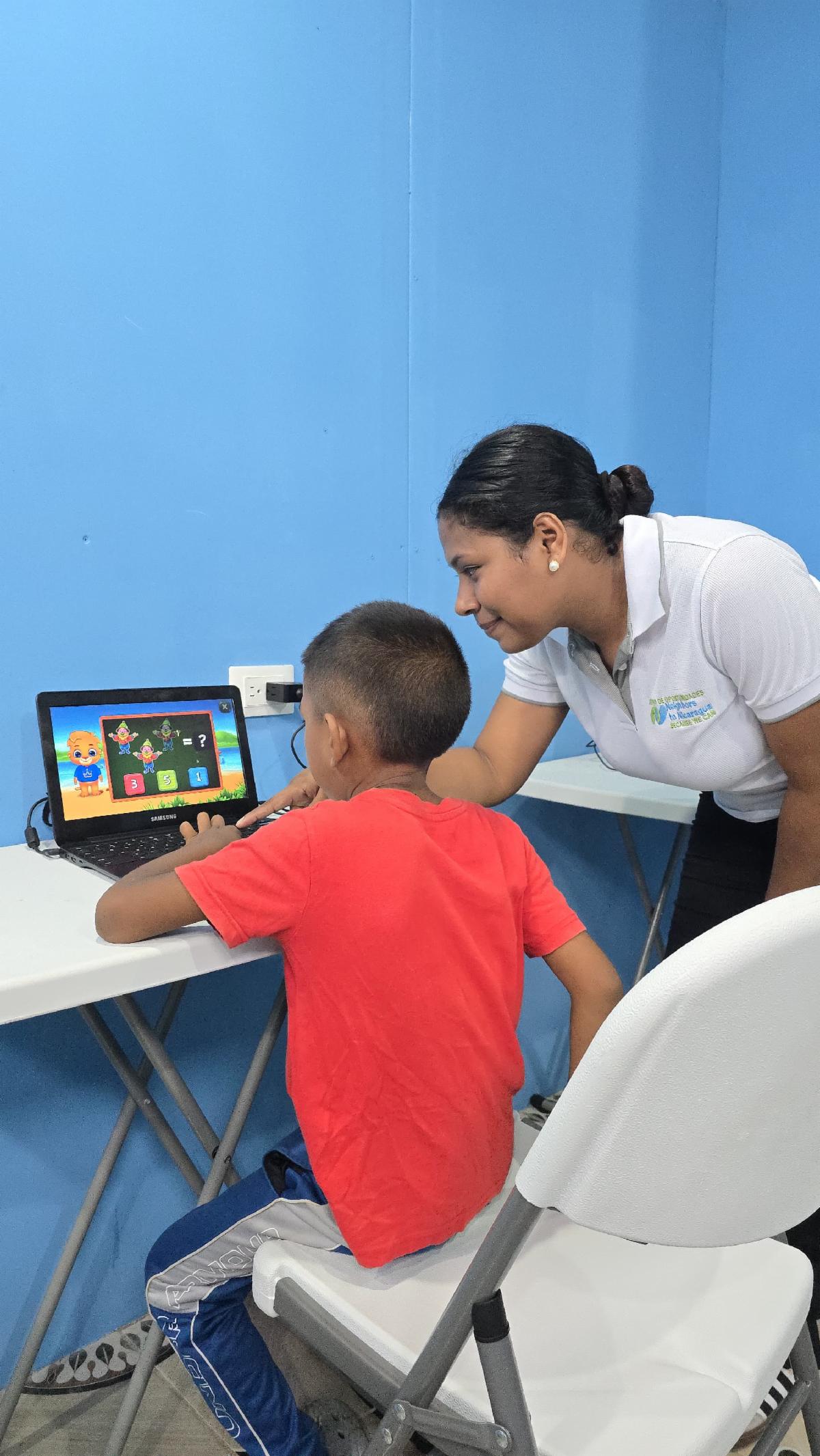 Teacher helps a boy as they use a tablet at a desk in a bright blue room.