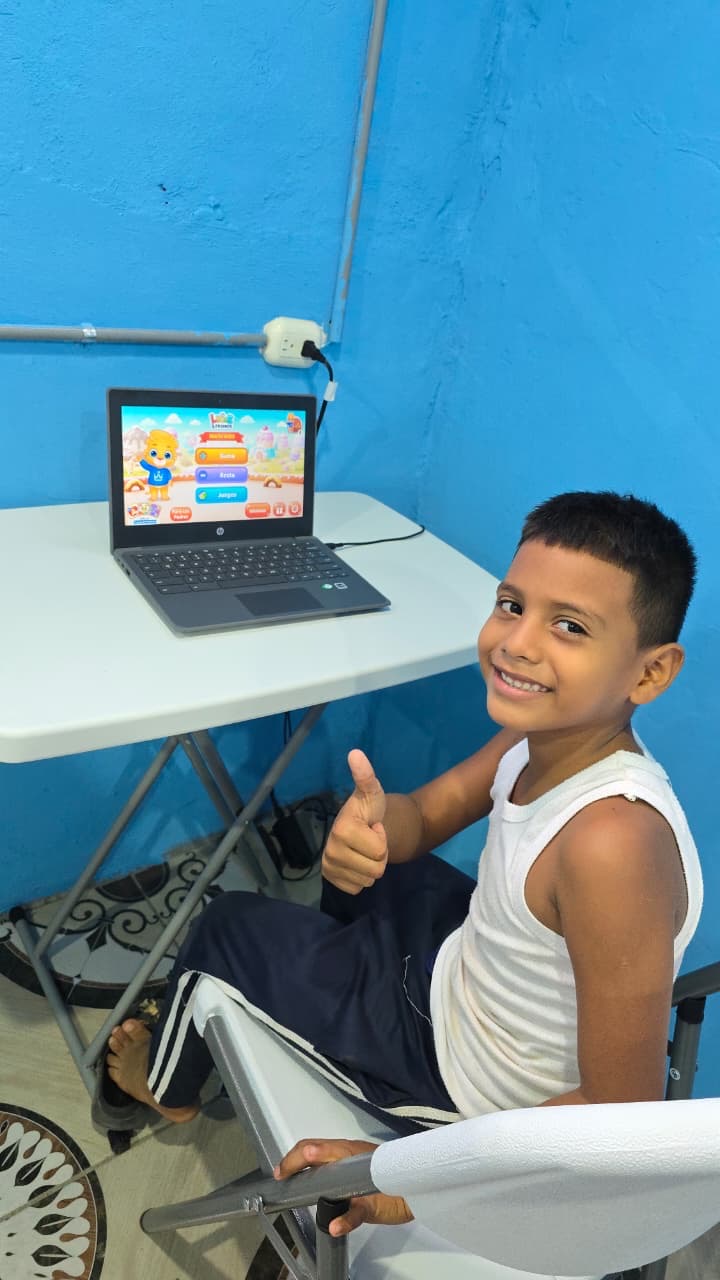 Smiling boy in a white tank top giving a thumbs up while using a laptop on a white table in a blue room