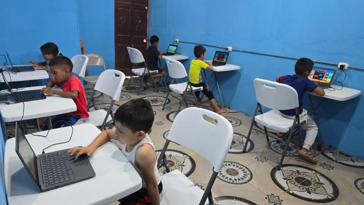 Children sit at individual desks in a bright blue computer lab, each using a laptop with wired headphones nearby (classroom learning).